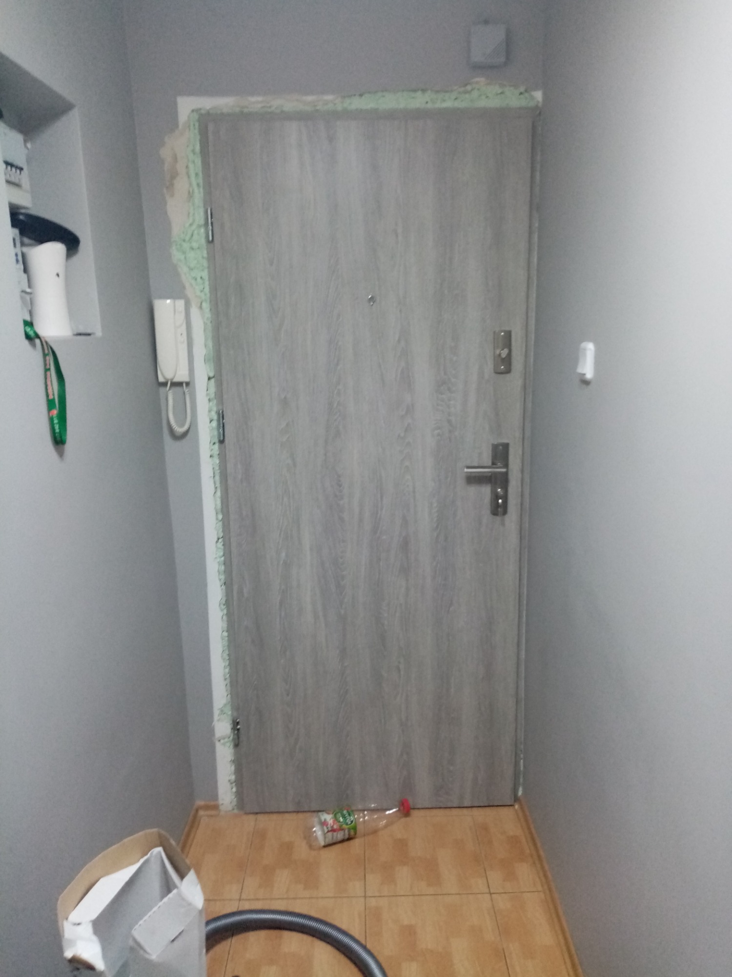 Montaż szarych drzwi wejściowych z ościeżnicą wypełnioną pianką montażową, widoczna butelka i karton na podłodze.