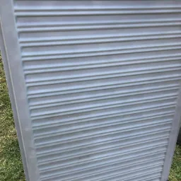 Szara, metalowa powierzchnia z poziomymi, równoległymi żłobieniami, prawdopodobnie po procesie piaskowania, leżąca na trawie.