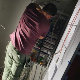 Elektro-instal - Elektryk w Chróścinie, w trakcie pracy nad rozbudowaną instalacją elektryczną w szafie rozdzielczej, widoczne liczne przewody i dokumentacja techniczna na ścianie.