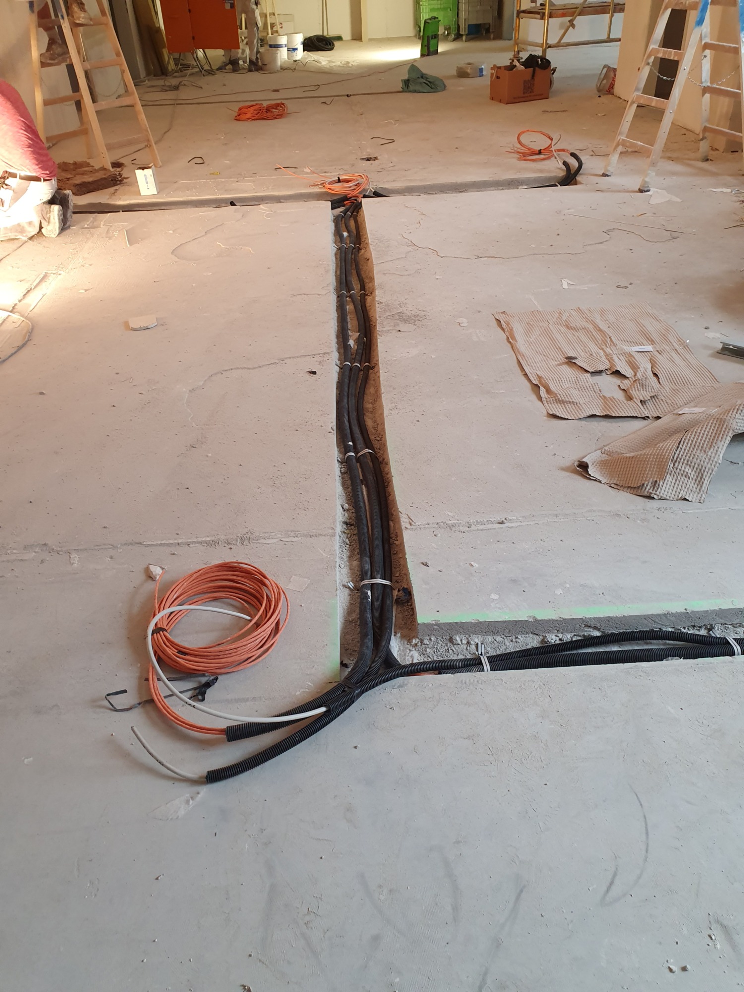 Wykop w surowej posadzce z ułożonymi przewodami elektrycznymi w peszlach, częściowo zabezpieczonymi białymi opaskami zaciskowymi, zwój pomarańczowego kabla obok, w tle drabiny i inne elementy...