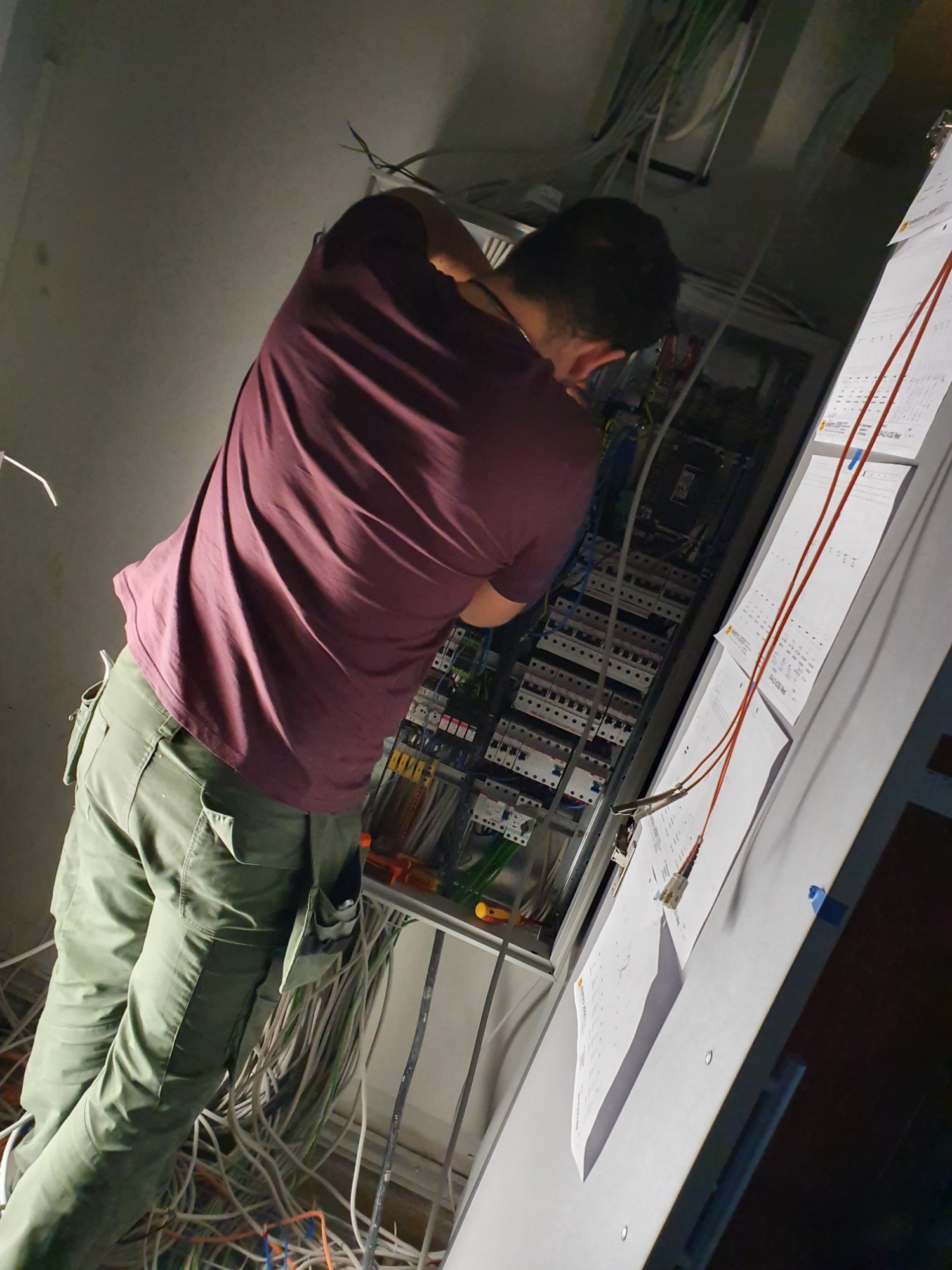 Elektryk w Chróścinie, w trakcie pracy nad rozbudowaną instalacją elektryczną w szafie rozdzielczej, widoczne liczne przewody i dokumentacja techniczna na ścianie.