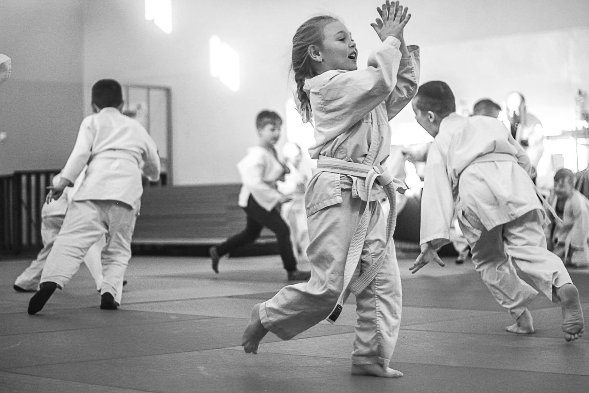 Czarno-białe zdjęcie grupy dzieci w białych strojach do sztuk walki, ćwiczących na macie w sali gimnastycznej, z jedną dziewczynką na pierwszym planie unoszącą ręce w geście radości.