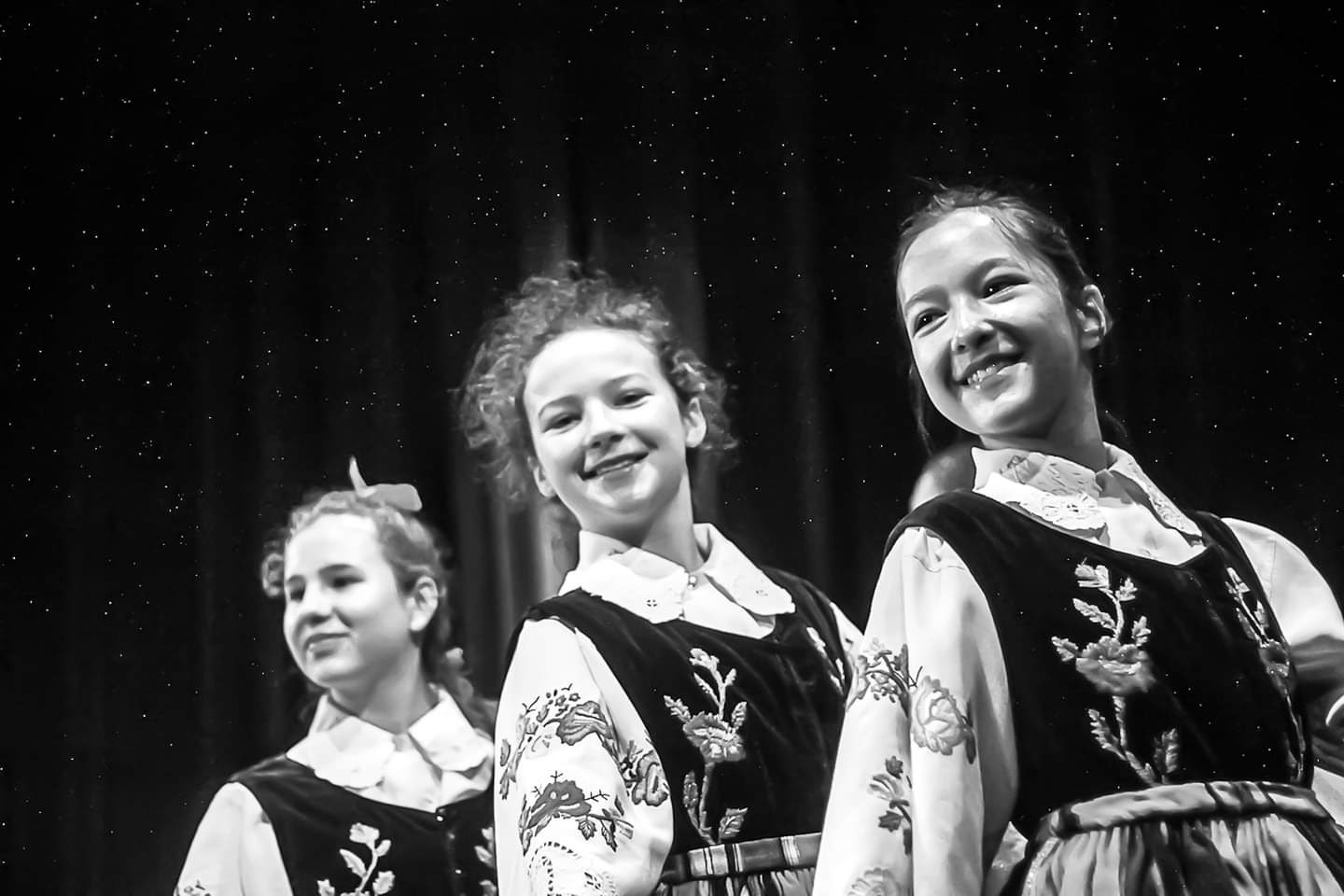 Trzy uśmiechnięte dziewczyny w ludowych strojach z haftowanymi kwiatami, na ciemnym tle przypominającym nocne niebo z gwiazdami, czarno-biała fotografia.