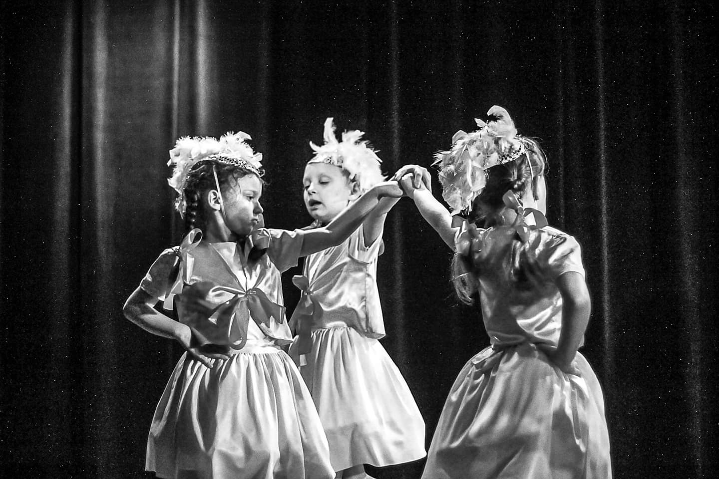 Czarno-białe zdjęcie trzech dziewczynek w kostiumach scenicznych z piórami na głowach, trzymających się za ręce na tle ciemnej kurtyny.