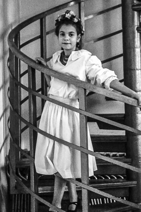 Dziewczynka w białej sukience i wianku na głowie stoi na krętych schodach, czarno-białe zdjęcie.