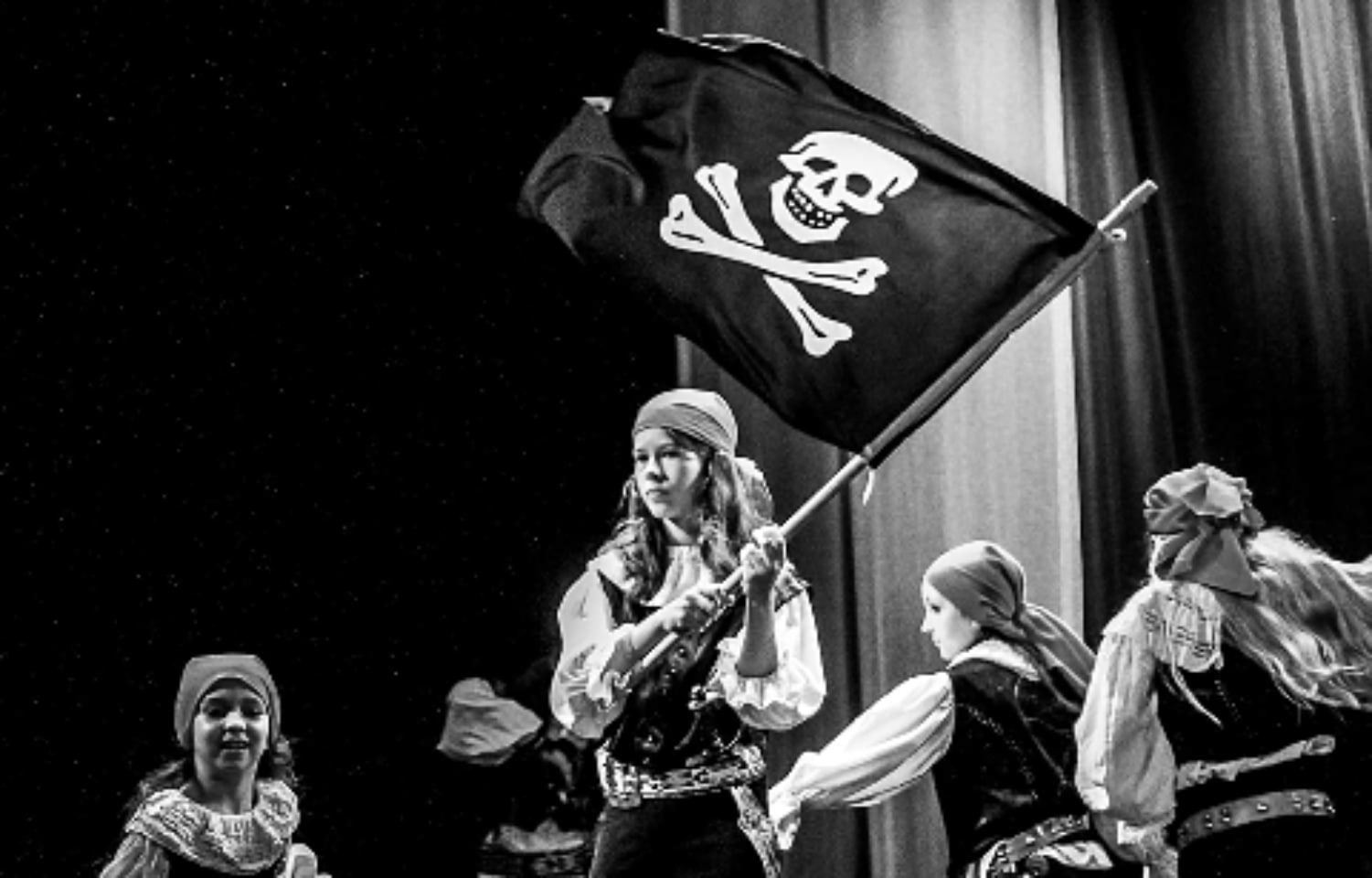 Czarno-białe zdjęcie grupy dzieci przebranych za piratów, w tym dziewczynki trzymającej flagę piracką z czaszką i skrzyżowanymi piszczelami na scenie podczas przedstawienia.