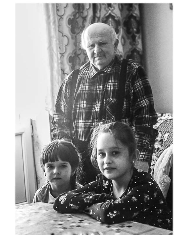 Czarno-białe zdjęcie przedstawia starszego mężczyznę w kraciastej koszuli i szelkach, stojącego za dwiema dziewczynkami siedzącymi przy stole. Dziewczynki mają ciemne włosy i patrzą w obiektyw. Tło...