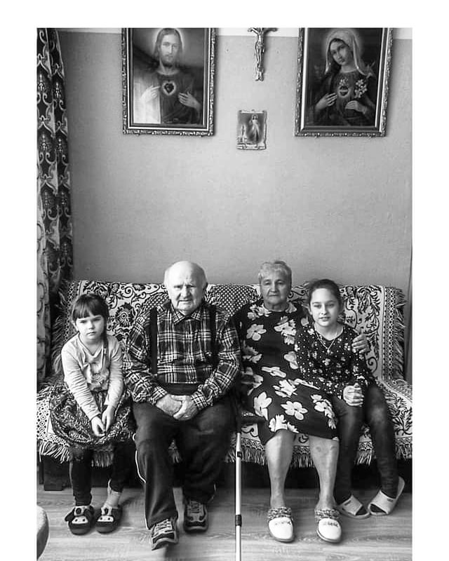 Czarno-białe zdjęcie przedstawia starsze małżeństwo siedzące na wzorzystej kanapie z dwiema dziewczynkami; nad nimi wiszą obrazy religijne, a obok kanapy widoczna jest laska.