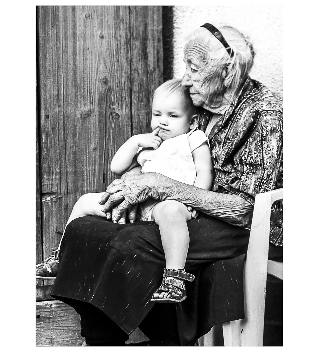 Czarno-biała fotografia przedstawia starszą kobietę siedzącą na krześle i trzymającą na kolanach małe dziecko, które wkłada palec do ust. W tle widoczne drewniane drzwi i ściana.