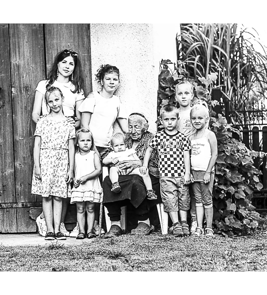 Czarno-białe zdjęcie portretowe wielopokoleniowej rodziny: babcia siedzi na krześle trzymając na kolanach małe dziecko, wokół niej stoi siedmioro dzieci i młodych dorosłych na tle drewnianych drzwi...