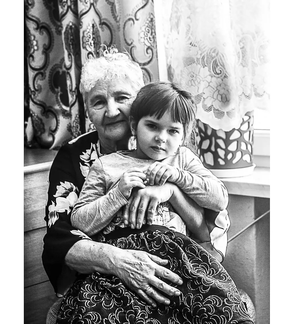 Czarno-białe zdjęcie przedstawia starszą kobietę z wnuczką na kolanach, siedzące na tle okna z firankami w kwiaty. Wnuczka ma poważny wyraz twarzy.