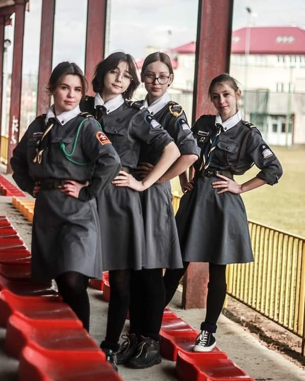 Cztery młode kobiety w szarych mundurkach harcerskich pozują na tle czerwonych trybun stadionu.