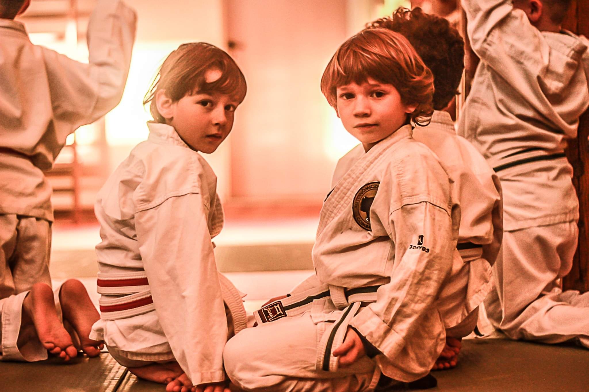 Grupa dzieci w białych kimonach siedzi na macie, czekając na zajęcia z karate, z widocznymi bose stopy i pasy z oznaczeniami stopni zaawansowania.