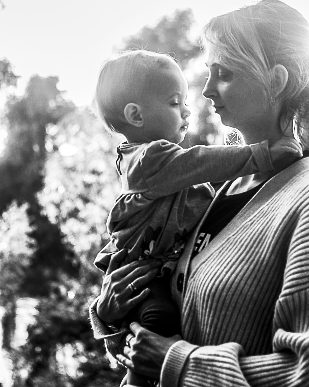 Czarno-białe zdjęcie matki trzymającej małe dziecko na rękach, oboje patrzą na siebie, delikatne światło pada z góry.