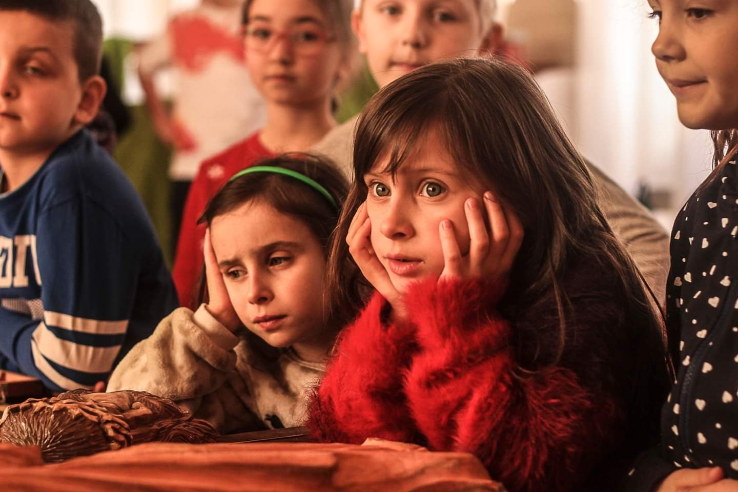 Grupa dzieci, w tym dwie dziewczynki z rękami podpartymi pod brodą, z zaciekawieniem obserwuje coś poza kadrem; jedna z dziewczynek ma na sobie czerwony sweter z futerkiem.