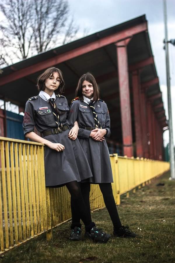 Dwie dziewczyny w szarych mundurkach harcerskich pozują oparte o żółte ogrodzenie na tle czerwonej konstrukcji zadaszenia.