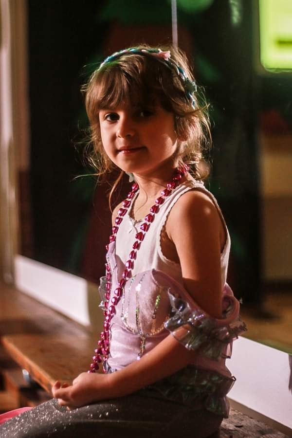 Portret uśmiechniętej dziewczynki w kostiumie z cekinami i różowymi koralami, siedzącej na drewnianej ławce z delikatnym oświetleniem