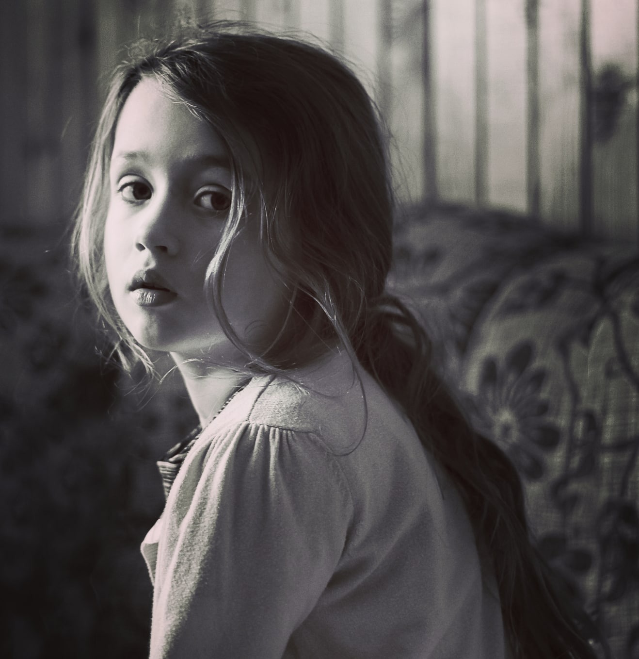 Portret dziewczynki z długimi włosami spiętymi w kucyk, odwróconej przez ramię, w czarno-białej tonacji, z widocznym wzorzystym obiciem sofy w tle.