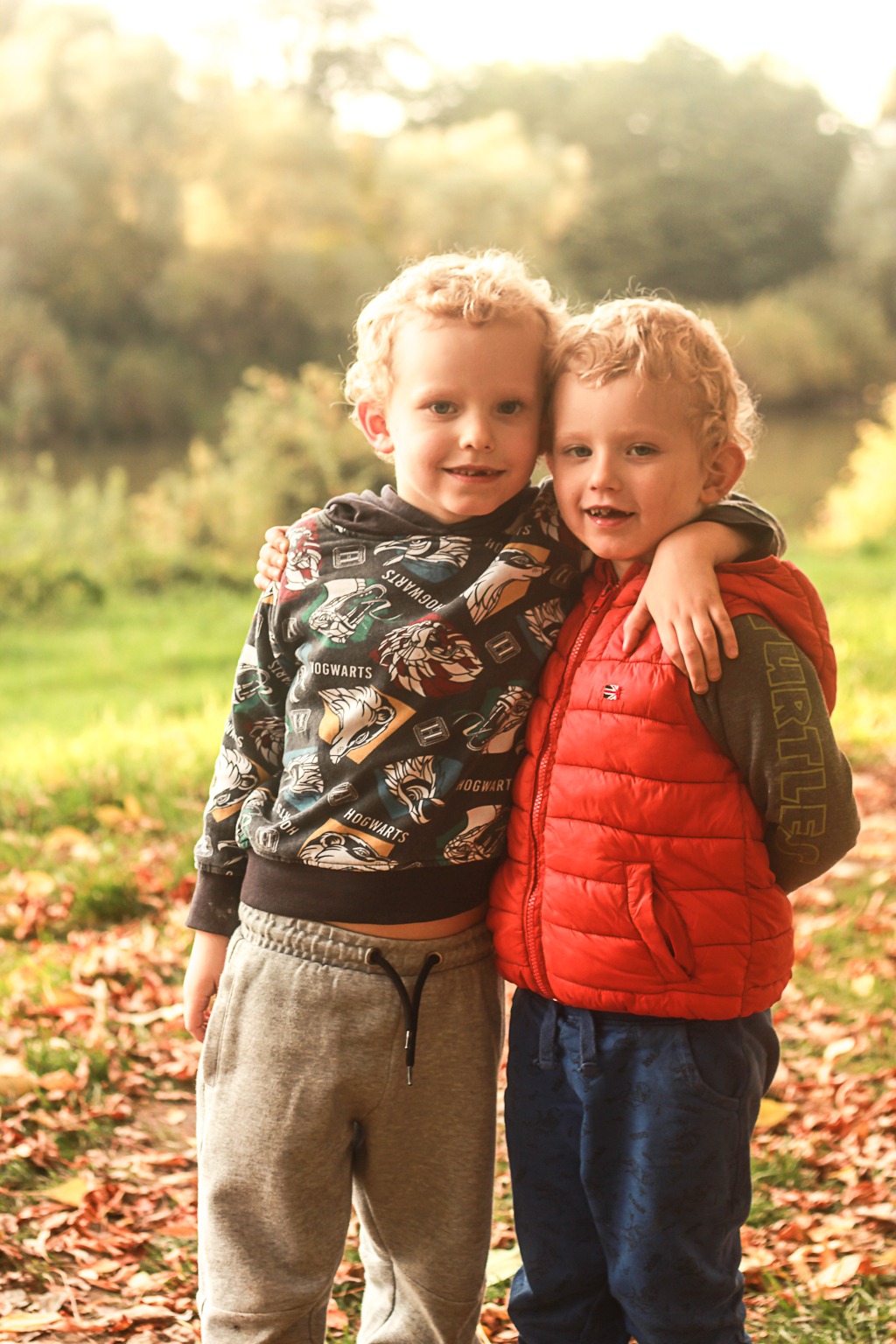 Dwóch blond chłopców obejmuje się na tle jesiennego parku, jeden w bluzie z motywem Hogwartu, drugi w czerwonej kamizelce.