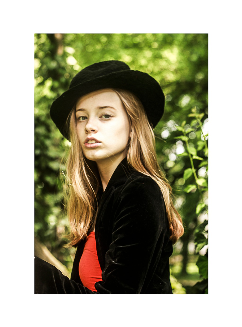 Portret młodej kobiety w czarnym kapeluszu i welurowej marynarce na tle zieleni, z delikatnym uśmiechem i spojrzeniem skierowanym w obiektyw.