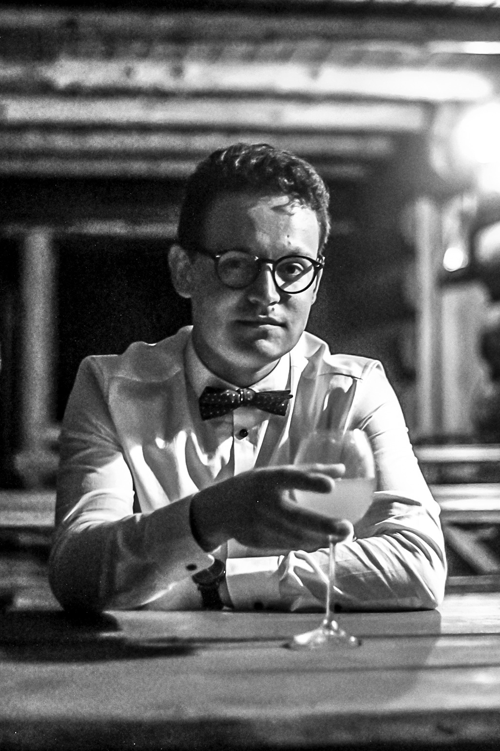 Mężczyzna w muszce i okularach, siedzący przy stole i trzymający kieliszek z jasnym napojem, zdjęcie czarno-białe.