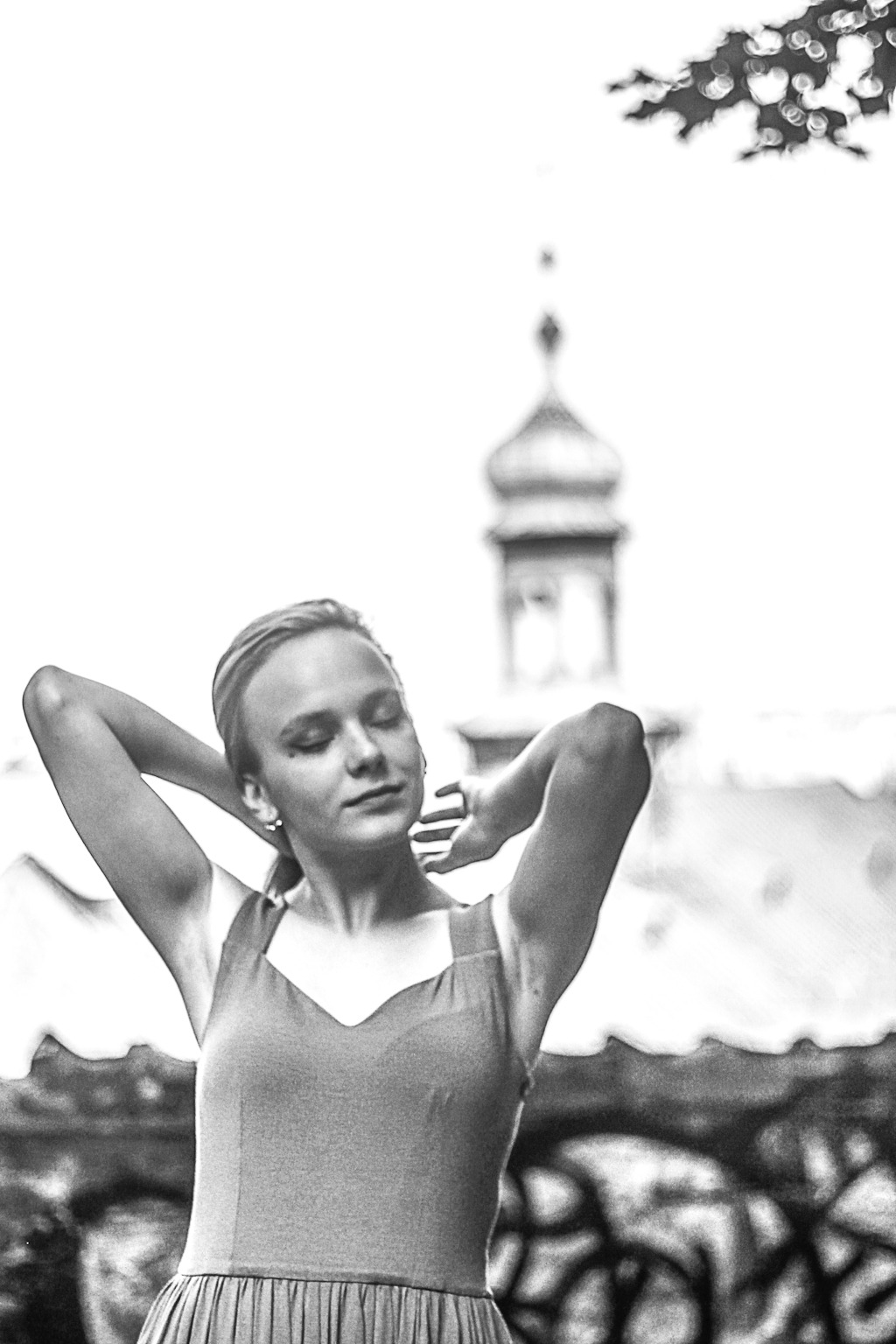 Kobieta w sukience z rękami uniesionymi za głowę, z zamkniętymi oczami, na tle rozmytego budynku z kopułą w czerni i bieli.