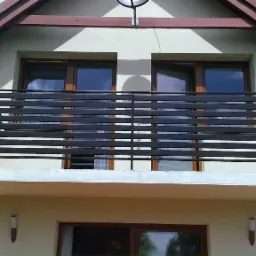 Nowoczesna balustrada balkonowa z poziomych, czarnych profili zamontowana na balkonie domu jednorodzinnego z kremową elewacją i brązowymi ramami okiennymi.