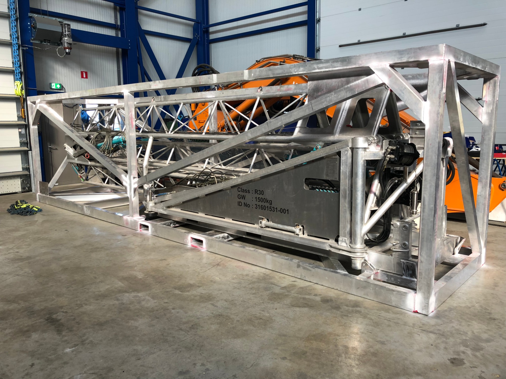 Metalowa rama konstrukcyjna z widocznym elementem hydraulicznym i tabliczką znamionową, na tle hali produkcyjnej z pomarańczowym elementem maszyny w tle.