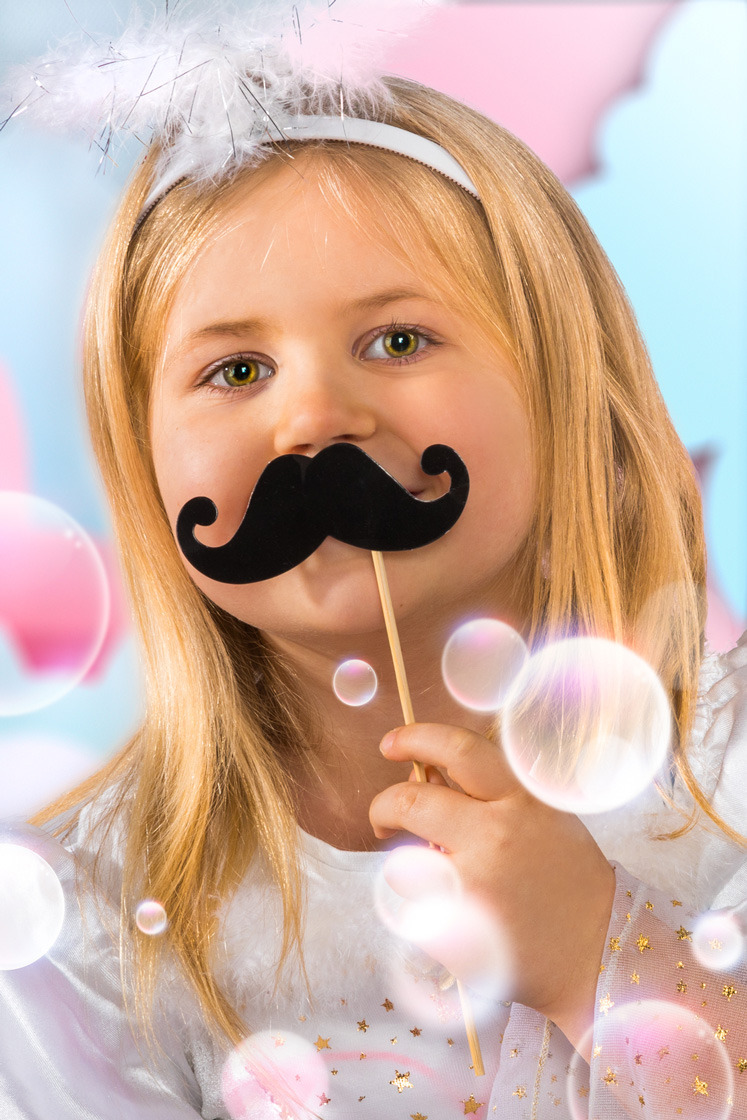 Dziewczynka z blond włosami i zielonymi oczami trzyma przed twarzą czarny, papierowy wąs na patyku, ubrana w białą bluzkę z gwiazdkami i opaskę z piórami, otoczona bańkami mydlanymi na jasnym tle.