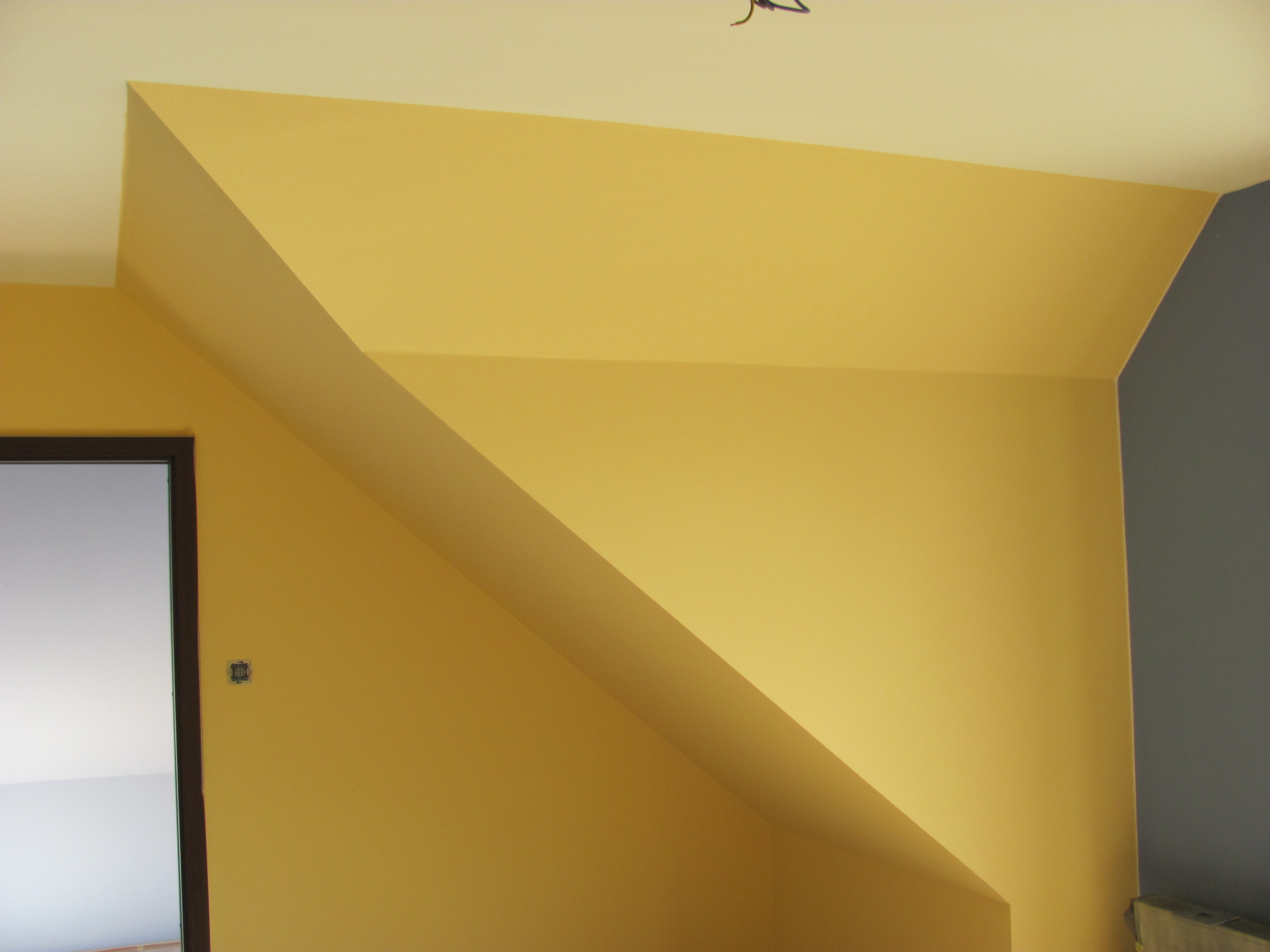Żółto-szare ściany na poddaszu po malowaniu, widoczna futryna drzwi i wystający element konstrukcyjny.