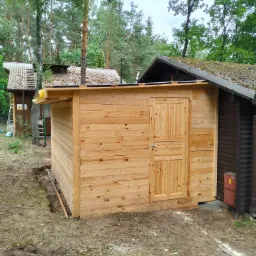 Świeżo postawiony, surowy drewniany budynek gospodarczy z drzwiami, widoczny na tle leśnej działki z innymi budynkami w tle.
