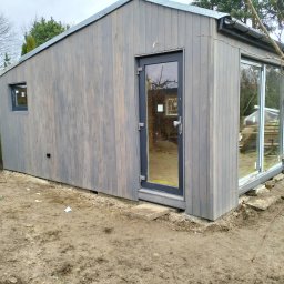 Alpinistyczna Wycinka Drzew - Nowoczesna, drewniana altana w odcieniach szarości z dużym oknem przesuwnym i drzwiami, umieszczona na nierównym, gliniastym terenie ogrodu.