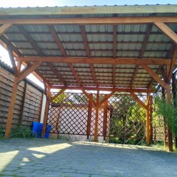 Alpinistyczna Wycinka Drzew - Drewniana konstrukcja z zadaszeniem z blachy falistej, częściowo osłonięta kratownicą, widziana z dołu na tle brukowanej nawierzchni i zieleni.