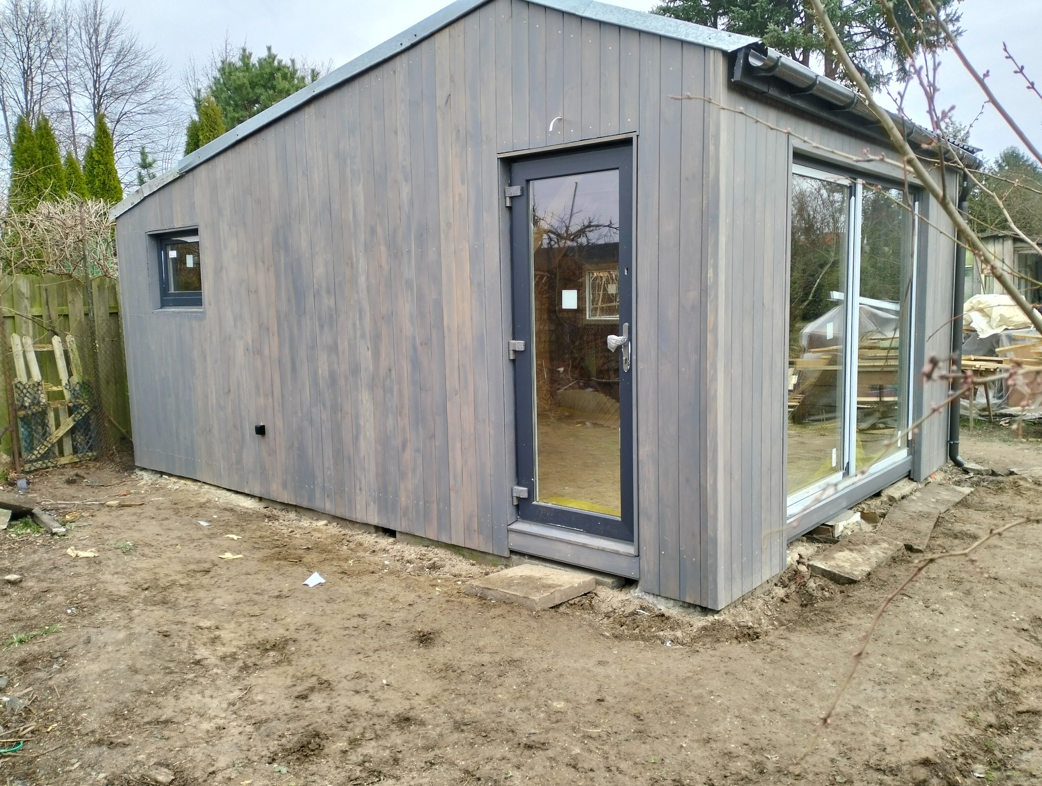 Nowoczesna, drewniana altana w odcieniach szarości z dużym oknem przesuwnym i drzwiami, umieszczona na nierównym, gliniastym terenie ogrodu.