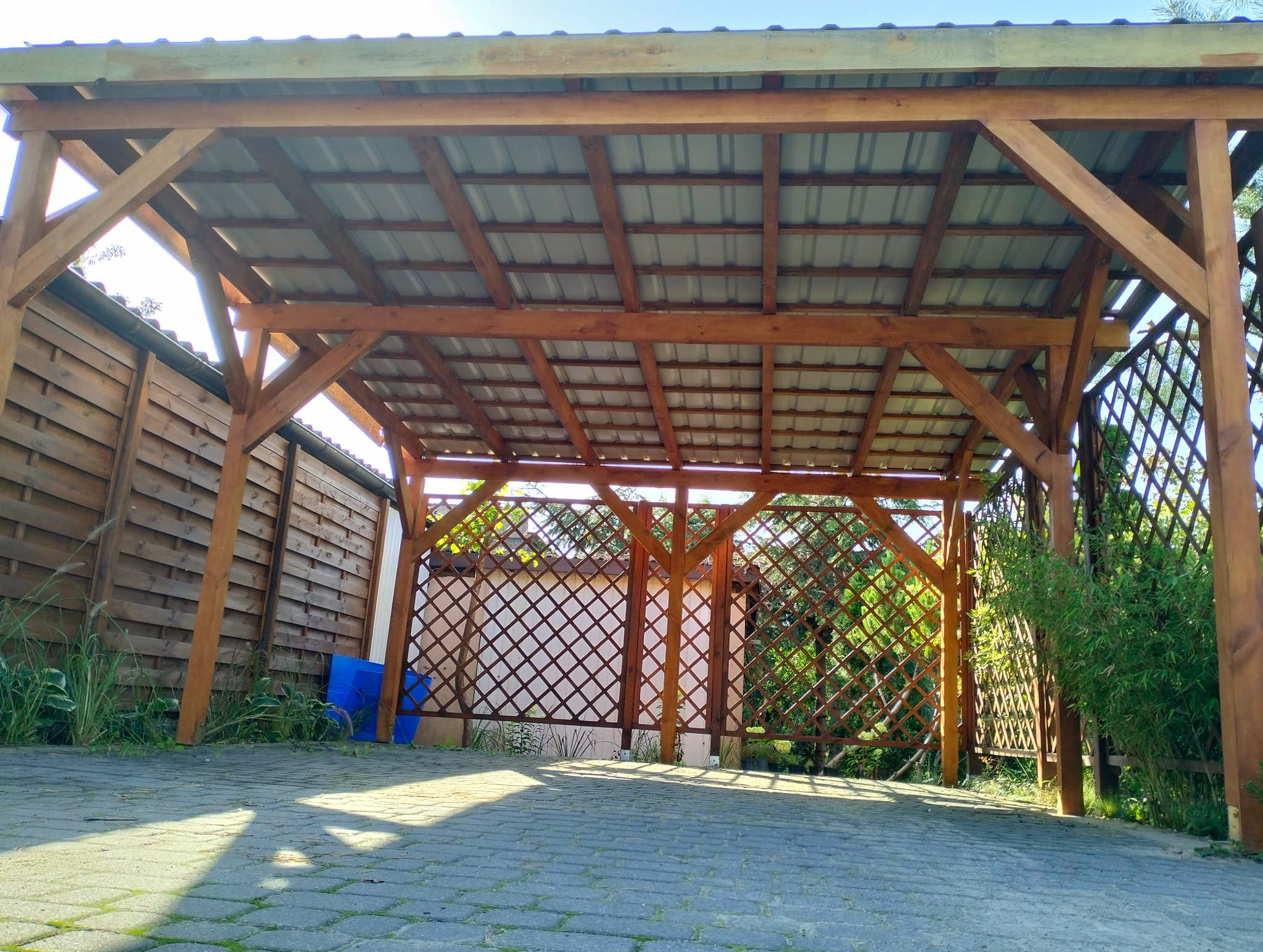 Drewniana konstrukcja z zadaszeniem z blachy falistej, częściowo osłonięta kratownicą, widziana z dołu na tle brukowanej nawierzchni i zieleni.