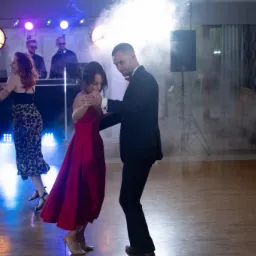 Para tańczy na parkiecie podczas wesela, w tle DJ, oświetlenie sceniczne i dekoracje stołu.