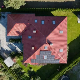 Widok z góry na dom z czerwoną dachówką, na której zamontowane są panele fotowoltaiczne. W tle zadbany ogród z równo przyciętymi żywopłotami i altaną.