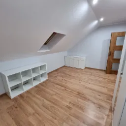 Poddasze po remoncie z oknem dachowym, białe ściany, drewnopodobna podłoga, biały regał z kwadratowymi przegródkami i biała szafka pod skosem.