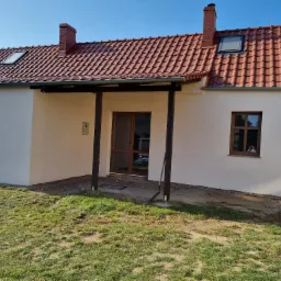 Dom jednorodzinny po renowacji elewacji i dachu z nową dachówką w odcieniach czerwieni oraz oknami dachowymi, widoczne świeże ocieplenie ścian zewnętrznych w jasnym odcieniu, zadaszenie tarasu...