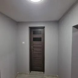 Szare wnętrze po odświeżeniu: ściany pomalowane na jednolity odcień szarości, drewnopodobna podłoga z szarych płytek, ciemne drzwi z przeszkleniami i okrągła lampa sufitowa.