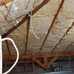 Izolacja poddasza wełną mineralną zamocowaną do drewnianych krokwi za pomocą metalowych uchwytów, widoczne przewody elektryczne, narzędzia DeWalt i metalowe profile na podłodze.