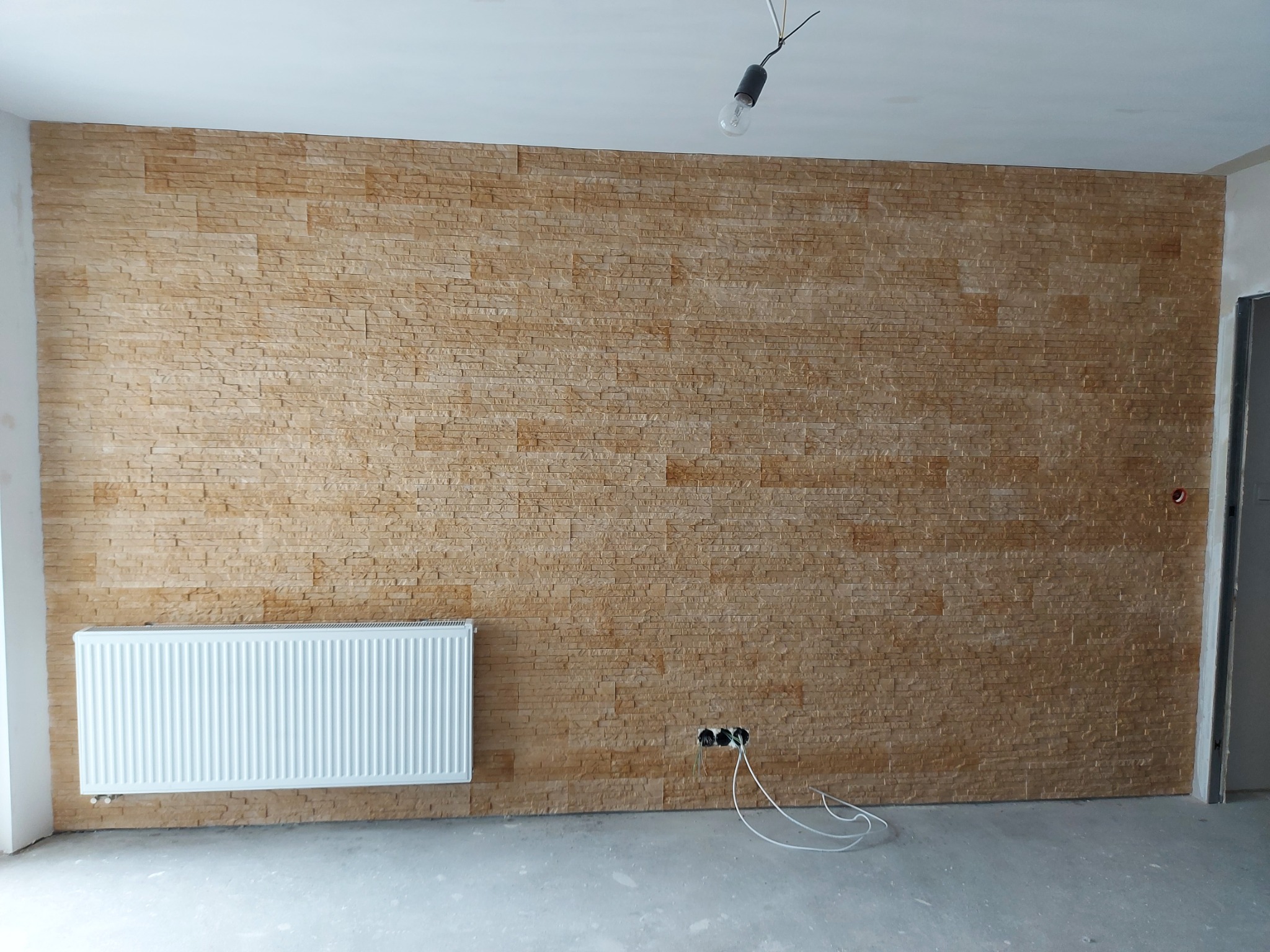 Ściana wewnętrzna wykończona panelami imitującymi kamień w odcieniach beżu i brązu, z zamontowanym białym grzejnikiem i wystającymi kablami elektrycznymi, surowy stan wykończenia wnętrza.