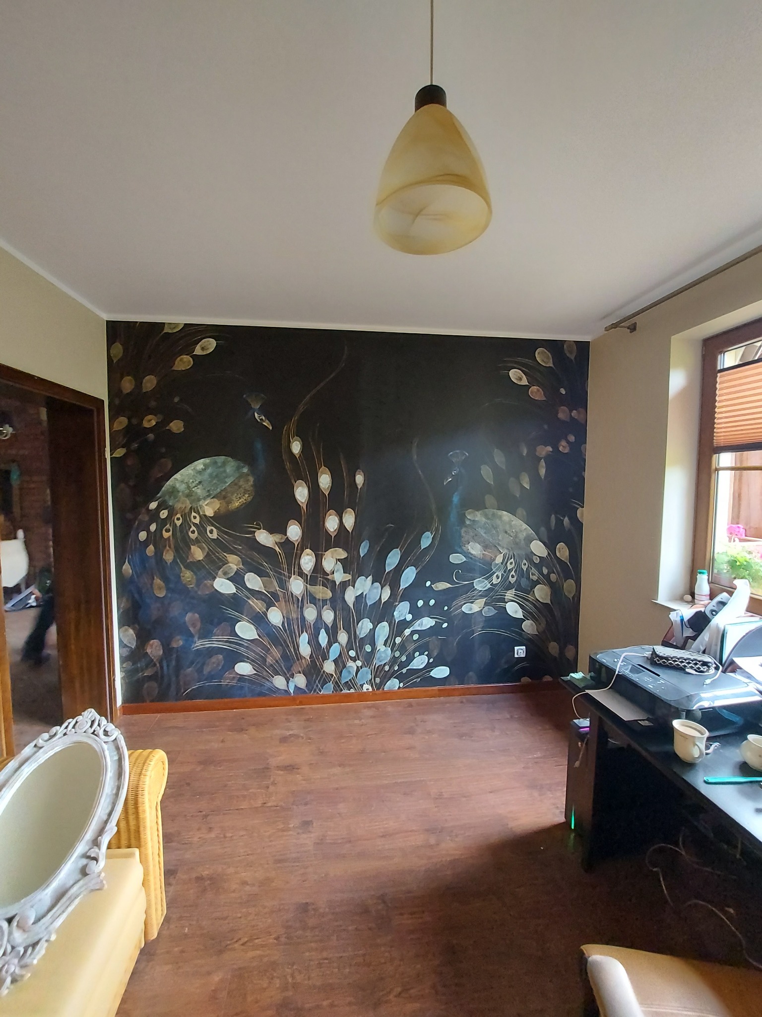 Pokój z ciemną ścianą ozdobioną wzorem pawich piór, drewnianą podłogą i lampą wiszącą. W tle widoczne biurko z akcesoriami i lustro w ozdobnej ramie.