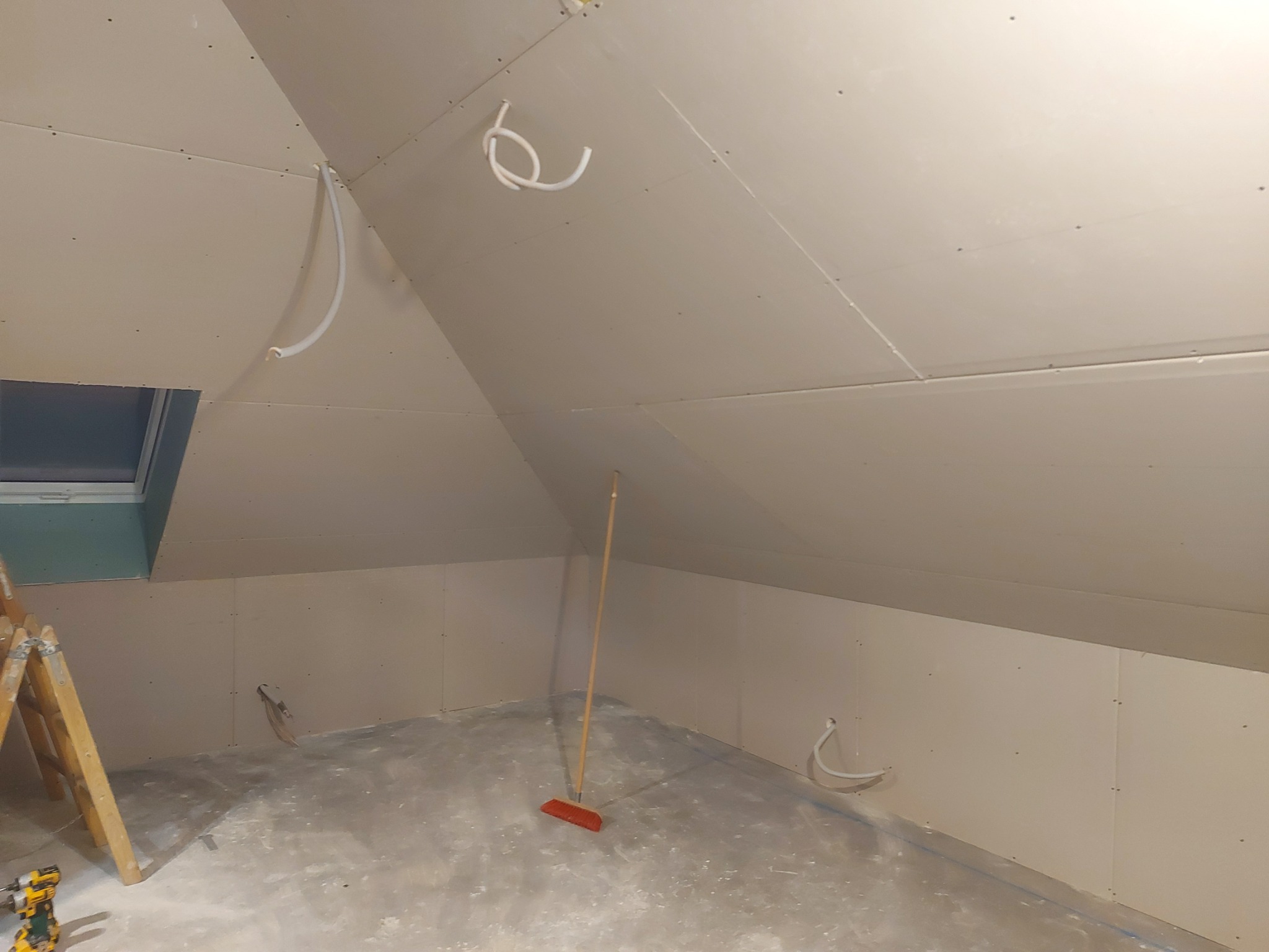 Poddasze w trakcie prac wykończeniowych: zamontowane płyty gipsowo-kartonowe na ścianach i skosach, widoczne wystające kable, drabina oparta o ścianę przy oknie dachowym, na podłodze leży miotła.