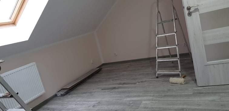 Poddasze po malowaniu ścian na jasny odcień, z widocznym oknem dachowym, grzejnikiem, drabiną i wałkiem do malowania na podłodze z paneli.