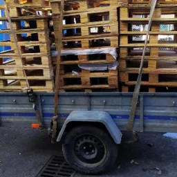 Przyczepa wypełniona drewnianymi paletami, zabezpieczonymi pasami transportowymi, stojąca na ulicy obok studzienki kanalizacyjnej.