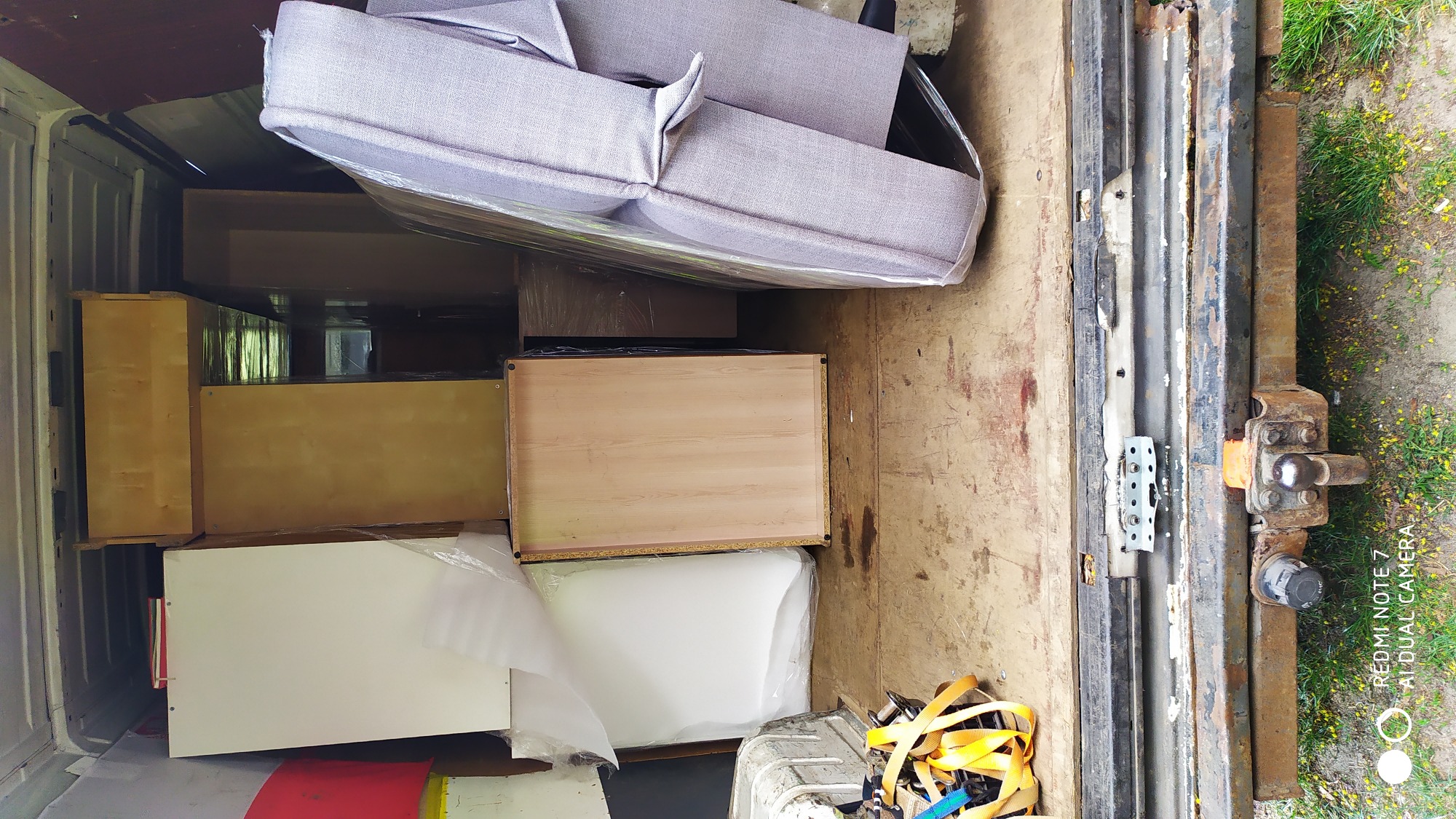 Wnętrze brudnej paki busa załadowane meblami, w tym szarą sofą owiniętą folią, drewnianymi szafkami i białymi elementami, zabezpieczone żółtymi pasami transportowymi.