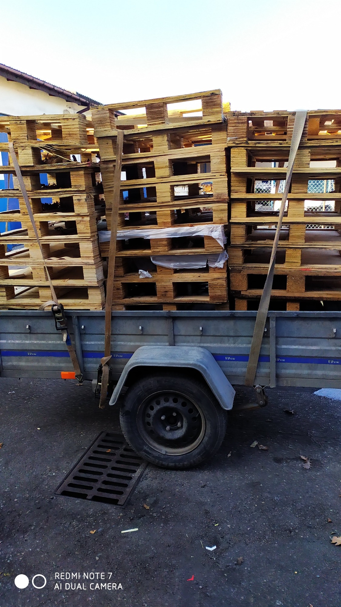 Przyczepa wypełniona drewnianymi paletami, zabezpieczonymi pasami transportowymi, stojąca na ulicy obok studzienki kanalizacyjnej.