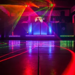 Parkiet oświetlony laserami w kolorach czerwonym i zielonym, w tle stanowisko DJ-a z ekranem i dodatkowym oświetleniem w kolorach niebieskim i fioletowym.