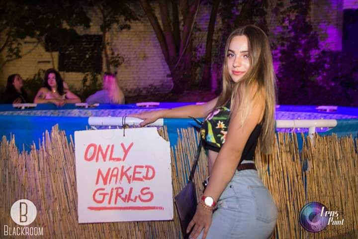 Młoda kobieta pozuje przed basenem z napisem 'Only Naked Girls' podczas wydarzenia plenerowego z oświetleniem dekoracyjnym.